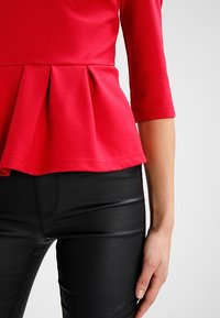 Kobieta w czerwonej, plisowanej bluzce w stylu peplum z rękawami do łokci oraz czarnych, dopasowanych spodniach, stojąca na tle jednolitego tła.