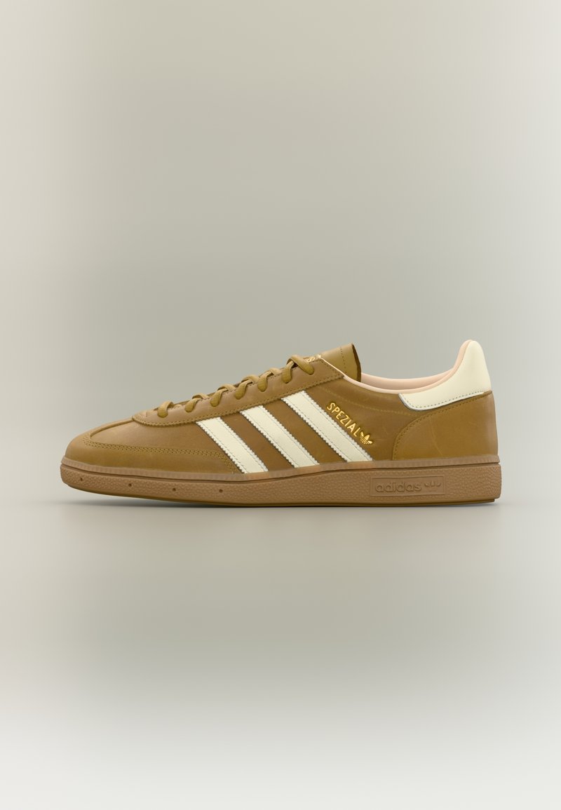 Καφέ αθλητικό παπούτσι Adidas Spezial με λευκές ρίγες, κρεμ ταμπελάκι στη φτέρνα και σόλα καραμέλας, εμφανιζόμενο σε πλευρική όψη πάνω σε ουδέτερο φόντο.