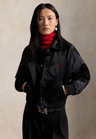 Frau mit langen dunklen Haaren trägt schwarze Satin-Bomberjacke mit rotem gesticktem Logo, rotem Rollkragenpullover und schwarzer Hose, steht mit den Händen in den Taschen.