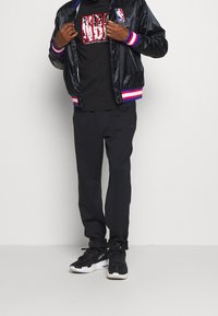 Černá saténová bomber bunda s červenými a modrými proužky na rukávech, černé tričko s grafikou NBA, černé kalhoty a černé tenisky s bílými detaily.