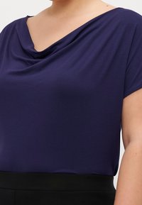 Marinblus med draperingar, gjord av mjukt tyg med vid halsringning och korta ärmar, i kombination med en svart midja längst ner.