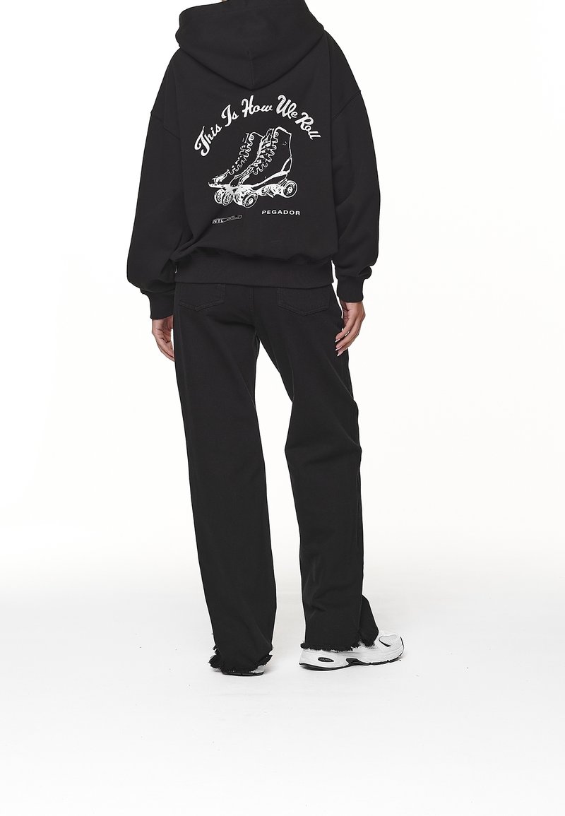 Personne portant un sweat à capuche noir avec un motif de patin à roulettes et le texte « This Is How We Roll », pantalon noir et baskets blanches, debout, de dos.