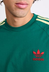 Zelena bombažna majica s kratkimi rokavi in krožnim izrezom, s pomočnimi rumenimi črtami na ramenih in rdečim vezenim logotipom Adidas na levem prsnem delu.