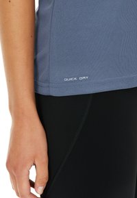 Grå hurtigttørrende træningsskjorte med en glat overflade, korte ærmer og en afrundet kant. Udstillet nær tætsiddende sorte leggings.