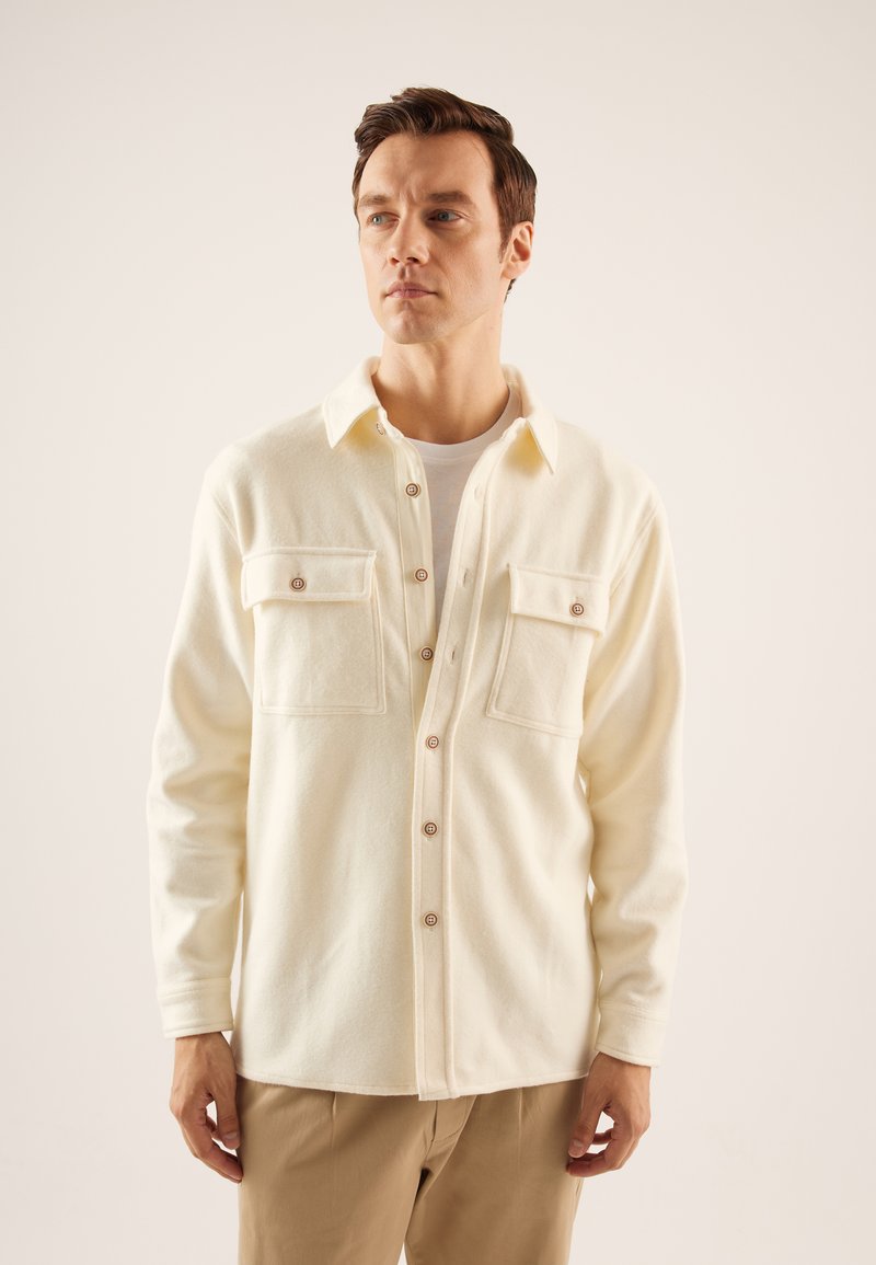 Cream-colored, button-up shirt jacket made of soft fabric; features two chest pockets, pointed collar, and wooden buttons.