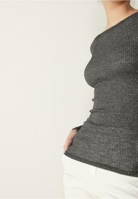 Kvinna i en figurnära, långärmad, mörkgrå ribbad tröja och vita byxor, med en hand i fickan, stående mot en enfärgad bakgrund.