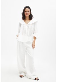 Blusa bianca con un ampio colletto da marinaio, dettagli in pizzo e maniche a volant, abbinata a pantaloni in lino bianchi a gamba larga e sandali neri.