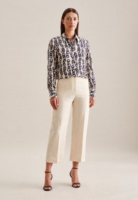 Camicia button-up a maniche lunghe con motivo a catena navy su tessuto crema, abbinata a pantaloni crema a vita alta e gamba larga, e sandali neri.