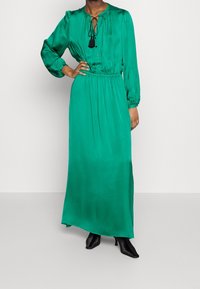 Lång klänning i smaragdgrön satin med långa ärmar, snörningshalsringning och elastisk midja. Svarta tofsdetaljer ger extra stil. Styld med svarta klackskor.