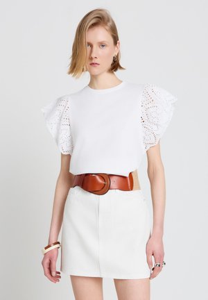 Femme portant un haut blanc avec des manches bouffantes à œillets, une jupe blanche, une large ceinture marron, et des bijoux minimalistes, debout devant un fond uni.