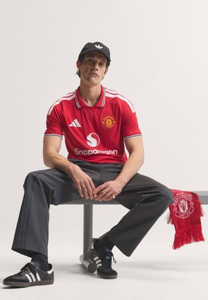 Mand iført en rød Manchester United fodboldtrøje og sort Adidas kasket sidder med et rødt halstørklæde over stolen mod en hvid baggrund.