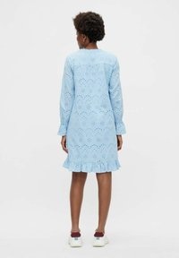 Moters su trumpais garbanotais plaukais, vilkinčios šviesiai mėlyną ilgomis rankovėmis nėriniuotą suknelę su volanų kraštu ir baltomis sportbačiais, stovinčios atsukusi nugarą.