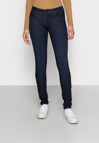 Jeans skinny azul oscuro con una textura suave, diseño de cinco bolsillos y sutiles detalles de costura, llevados con zapatillas blancas.