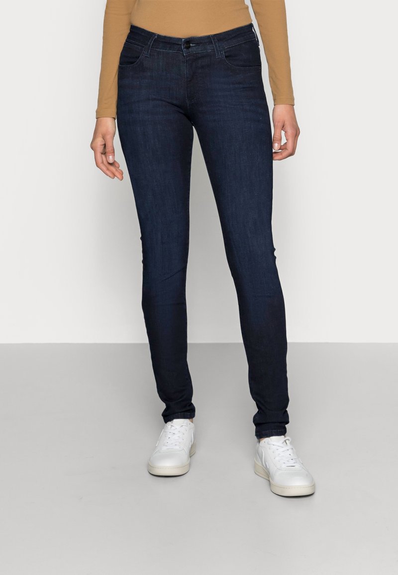 Jeans skinny azul oscuro con una textura suave, diseño de cinco bolsillos y sutiles detalles de costura, llevados con zapatillas blancas.