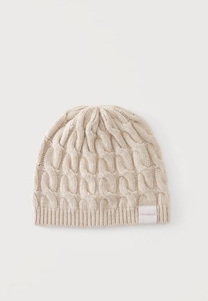 Beige strikket hue med chunky kabelstrikmønster og ribbet kant, med et lille hvidt mærke med rød tekst på den ene side.