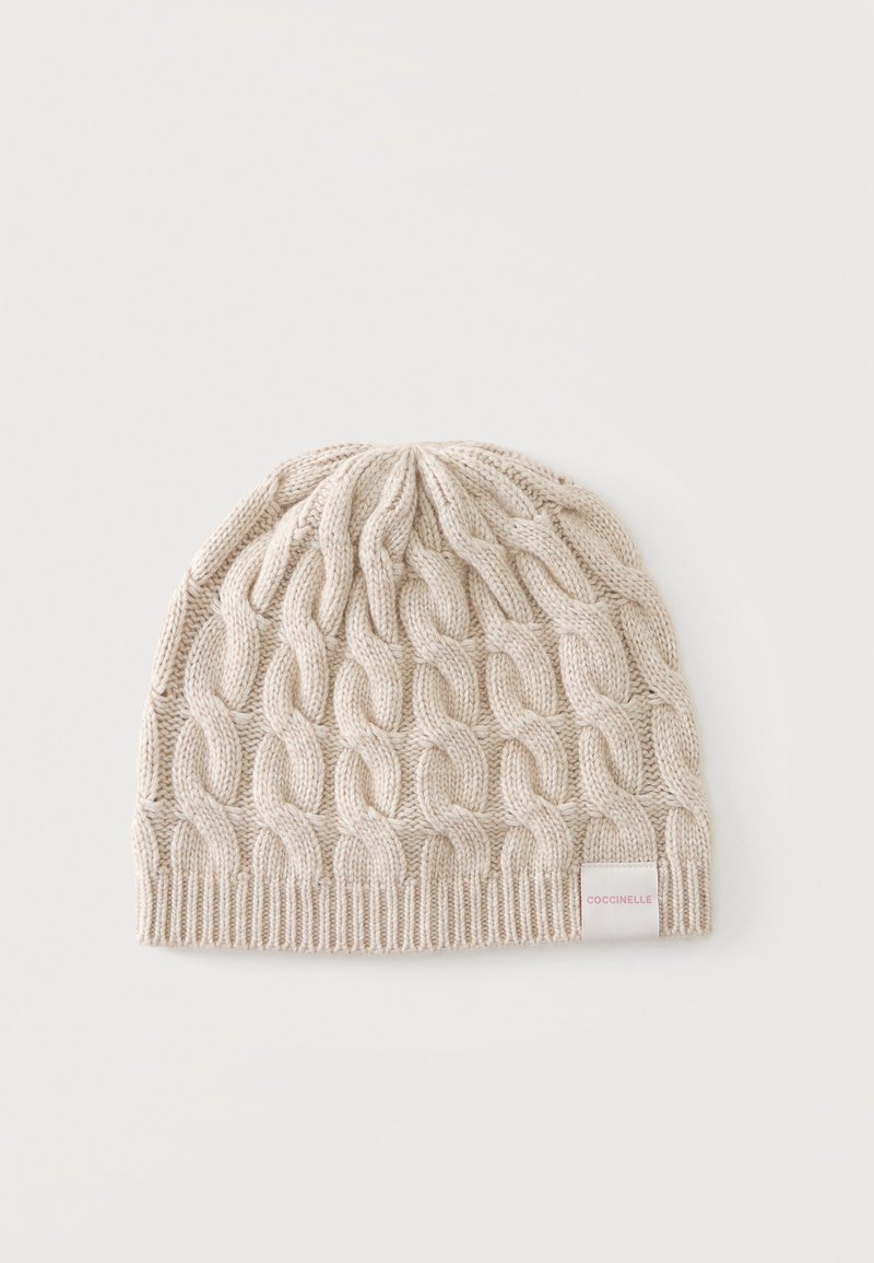 Bonnet beige tricoté avec un motif en grosse torsade et une bordure côtelée, avec une petite étiquette blanche avec un texte rouge sur un côté.