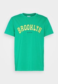 Camiseta de algodón verde de manga corta con texto amarillo "BROOKLYN" en arco sobre el pecho y pequeño logo negro "KANGOL" debajo.