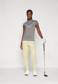 Šedé golfové polo s límcem na zip a geometrickým vzorem, doplněné světle žlutými kalhotami. Dokončeno bílými botami a golfovou holí v ruce.