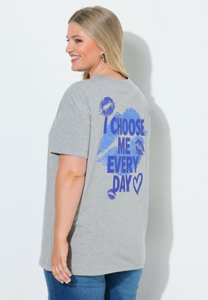 Donna sorridente con lunghi capelli biondi indossa una t-shirt grigia con testo blu e segni di baci che dicono "I choose me every day" e un simbolo a forma di cuore.