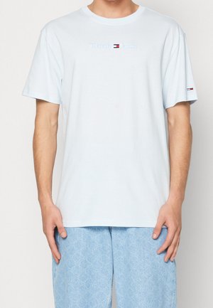 Mężczyzna noszący jasnoniebieską koszulkę Tommy Jeans z logo na klatce piersiowej i rękawie, w połączeniu z jasnoniebieskimi spodniami w wzory.
