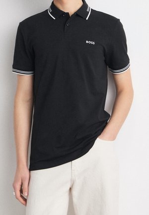 Schwarzes Polohemd mit kurzen Ärmeln, weißen Doppelstreifen am Kragen und an den Bündchen und kleinem weißen "BOSS"-Logo auf der Brust, getragen zu hellen Hosen.