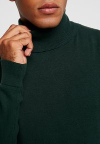 Uomo che regola un maglione a collo alto rigato verde scuro, mostrando un primo piano della mano, del collo e della parte superiore del torso.