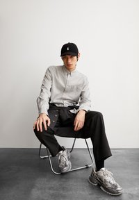 Lichtgrijze button-up shirt met zak, zwarte baseballpet met een witte band, zwarte broek met knoopdetails en grijze sneakers.