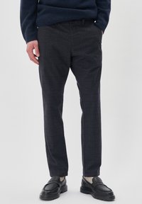 Uomo che indossa pantaloni a quadri grigio scuro, maglione blu navy, calzini beige e mocassini in pelle nera, in piedi con una mano in tasca.