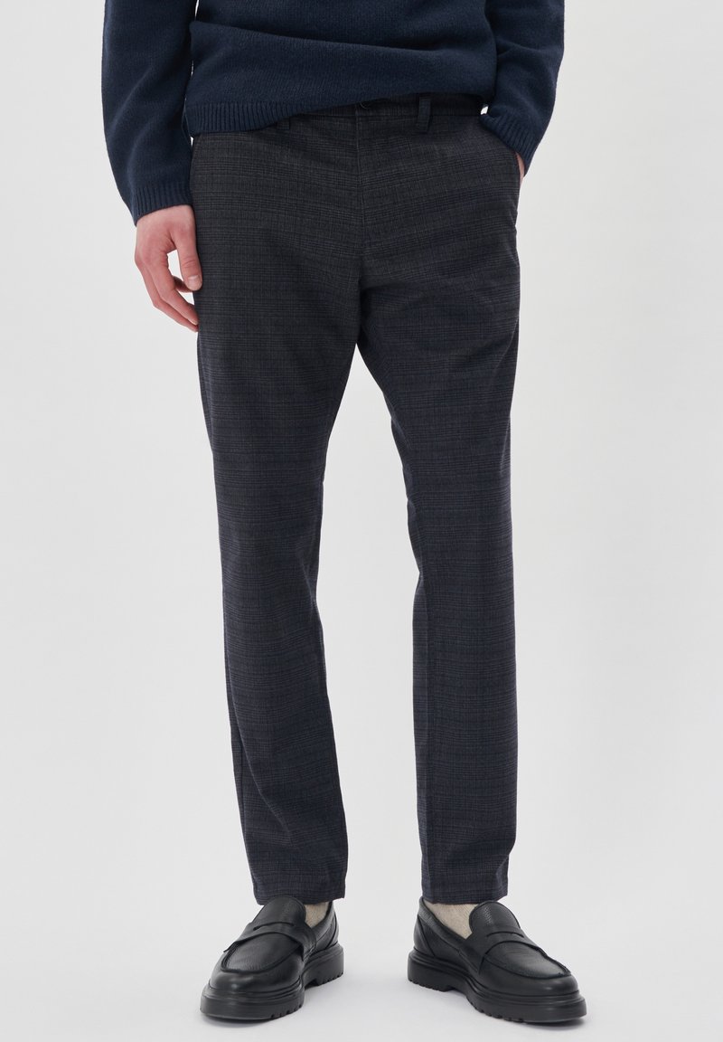 Uomo che indossa pantaloni a quadri grigio scuro, maglione blu navy, calzini beige e mocassini in pelle nera, in piedi con una mano in tasca.
