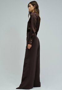 Mujer de perfil vistiendo una blusa de seda marrón oscuro de manga larga metida en unos pantalones de pierna ancha de tiro alto con zapatos de punta.