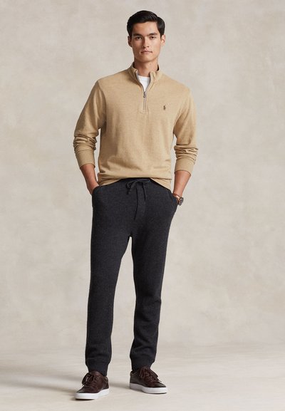 Moški stoji z rokami v žepih, oblečen v bež pulover s četrtinskim zadrgo, temno sive hlače z vrvico in temno rjave superge na nevtralnem ozadju.