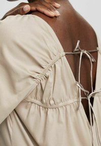 Personne à la peau foncée portant une robe beige en tissu froncé avec un dos ouvert noué par deux nœuds, main touchant l'arrière de leur cou.