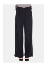 Pantaloni neri a gamba larga con vita alta, caratterizzati da pieghe, chiusura con bottone e una texture liscia. Indossati con scarpe nere con punta appuntita.