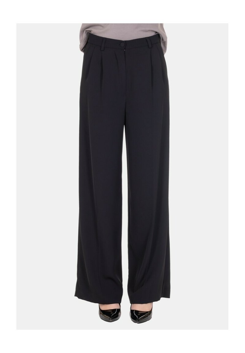 Pantaloni neri a gamba larga con vita alta, caratterizzati da pieghe, chiusura con bottone e una texture liscia. Indossati con scarpe nere con punta appuntita.