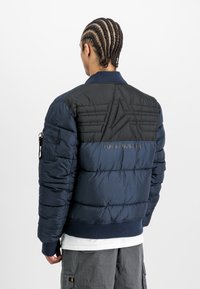 Moški z pletenimi lasmi, oblečen v modro-črno prešito jakno znamke Alpha Industries, s sivimi cargo hlačami, obrnjeno proti prazni beli ozadju.