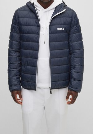 Persoon draagt een donkerblauwe gewatteerde jas met "BOSS"-logo over een witte hoodie en bijpassende witte broek, staand tegen een effen achtergrond.