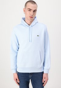 Sudadera azul clara hecha de tela suave, con un bolsillo frontal, capucha con cordón y un pequeño logo de Lacoste en el pecho.