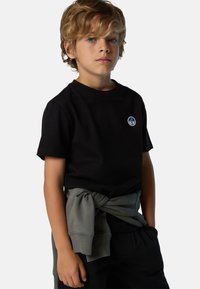 Camiseta de algodón negra con un pequeño logotipo circular azul, combinada con pantalones oscuros y una sudadera gris atada a la cintura. Mangas cortas.