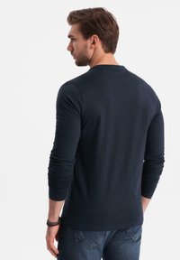 Hombre vestido con una camiseta de manga larga oscura y jeans azules, de pie con la espalda vuelta y mirando hacia la izquierda contra un fondo sencillo.
