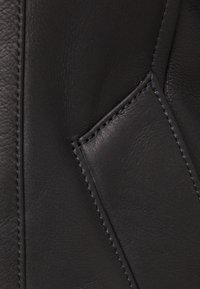 Detailní záběr na černou kůži s hladkým povrchem, který vyznačuje výrazné švy a malou kapsu. Je patrná bohatá, tmavá barevnost.