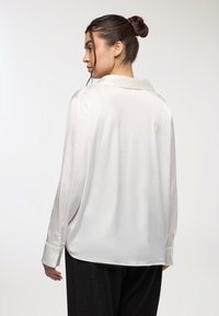 Donna con i capelli raccolti a chignon, che indossa una camicia bianca di satin larga e pantaloni neri, voltata di spalle con la testa girata di lato.