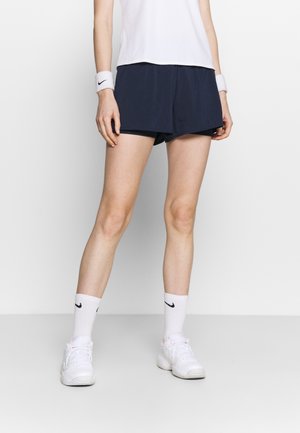Personne portant un short de sport noir, un haut de sport blanc, des poignets Nike blancs, des chaussettes et des baskets blanches sur un fond uni.