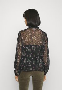 Blusa nera trasparente con motivo floreale in rosa e verde, maniche lunghe, dettagli a pois e scollatura posteriore con bottoni.