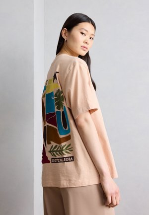 Γυναίκα με μακριά σκούρα μαλλιά που φοράει μια μπεζ φαρδιά μπλούζα με πολύχρωμο αφηρημένο σχέδιο και κείμενο "SCOTCH&SODA" στην πλάτη.