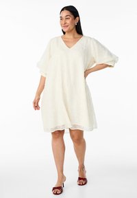 Robe couleur crème, longueur genou, avec un décolleté en V et des manches courtes bouffantes, fabriquée en tissu texturé. Associée à des sandales à talons rouges.
