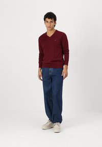 Bordeaux V-hals trui van zachte stof, gecombineerd met losse blauwe jeans en lichtgekleurde schoenen. Minimale merkdetails zichtbaar.