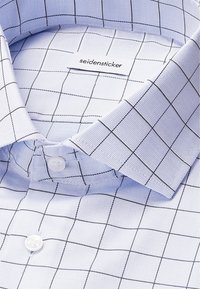 Camicia a scacchi blu chiaro con colletto strutturato, bottoni bianchi e un tessuto testurizzato con motivi a griglia nera. Etichetta del marchio visibile.