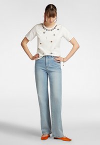 Camicetta bianca a maniche corte con ricami floreali, abbinata a jeans a gamba larga azzurri e ballerine arancioni. Design semplice, vestibilità rilassata.