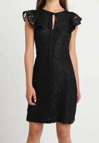 Svart spetsklänning med korta, volangärmar, en nyckelhålsknäppning med knytning och en figurnära silhuett, med blommönster över hela.