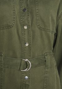 Oliwkowa kurtka z teksturowanej tkaniny, z metalowymi zatrzaskami, dwoma kieszeniami na piersi oraz paskiem materiałowym z metalowym detalem w postaci okręgu.
